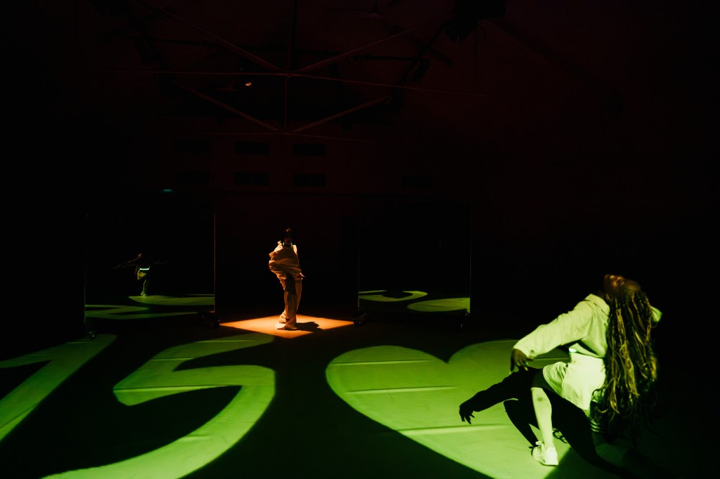 Two dancers are on a white dance floor. One is in the back of the room in a lit up diamond. Another is in the front right foreground leaning over. The words 15-Love
